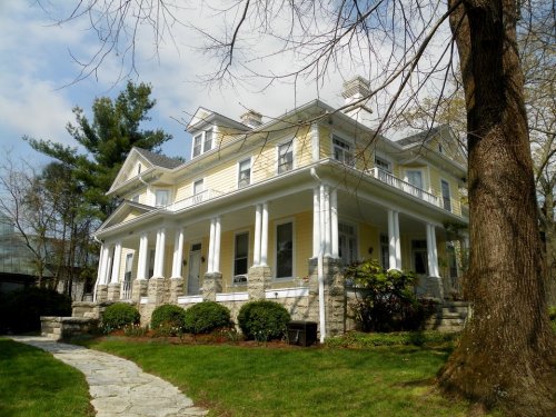 A large yellow house with white columns, surrounded by greenery and a stone pathway, under a partly cloudy sky.