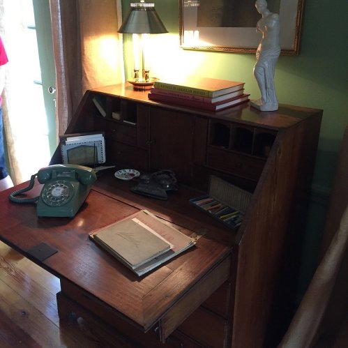 A wooden desk with a vintage phone, books, and a lamp, beside a statue and a window with curtains.