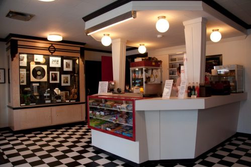 A retro-style diner interior with a checkered floor, a service counter, and display cases filled with snacks and memorabilia.