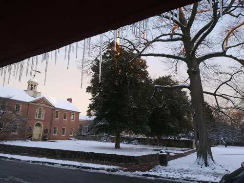 A snowy landscape featuring a historic building, bare trees, and icicles hanging from a roof.
