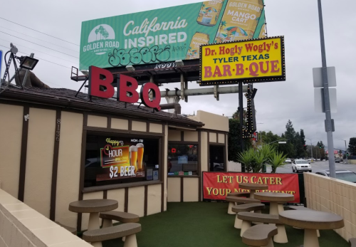 A BBQ restaurant with outdoor seating, a large sign, and a billboard above advertising California-inspired food.