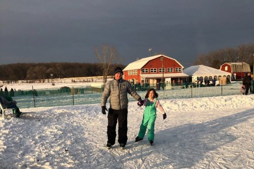A man and a girl skate on an outdoor rink, surrounded by snow and a red barn in the background.