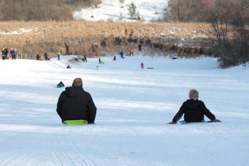 Two people sit on sleds at the top of a snowy hill, with others sledding down in the background.
