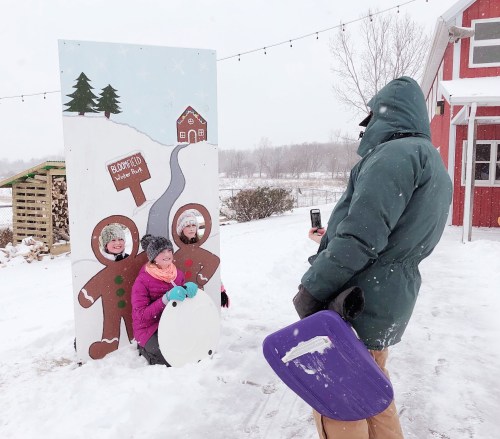 Two children in winter gear pose behind a snowy backdrop, while an adult takes a photo with a phone. Snow is falling.