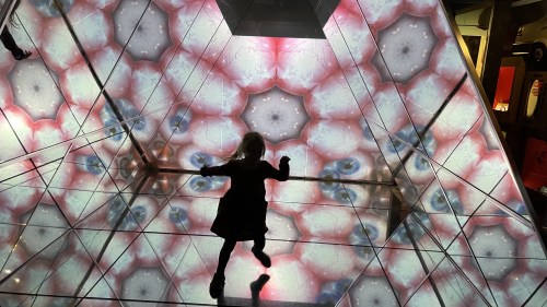 A child plays inside a kaleidoscopic installation with colorful patterns reflecting on the walls and floor.
