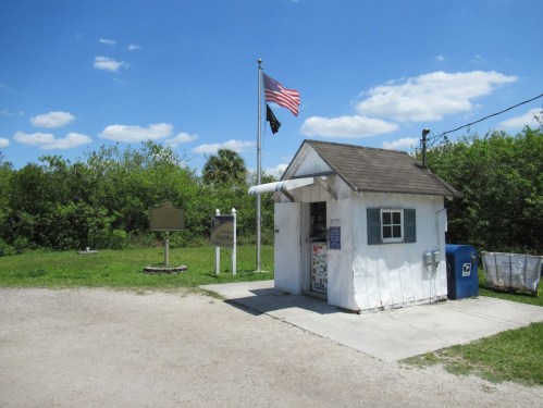 A small white building with a gray roof, American flag, and greenery in the background under a blue sky.
