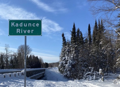 Sign for Kadunce River beside a snowy landscape with evergreen trees under a clear blue sky.