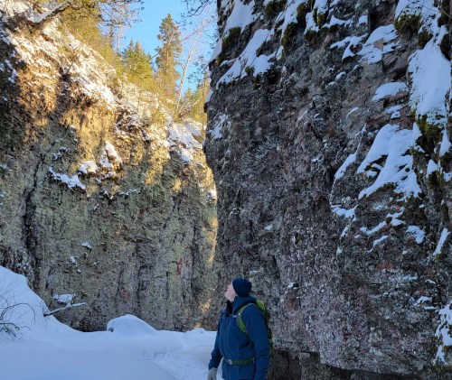 A person in winter attire gazes up at towering rock formations in a snowy landscape. Sunlight filters through the trees.