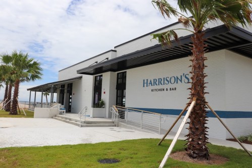 Exterior view of Harrison's Kitchen & Bar, featuring palm trees and a modern design by the waterfront.