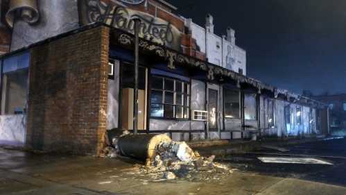 A damaged building with graffiti, featuring a broken column on the sidewalk, illuminated by nighttime lighting.