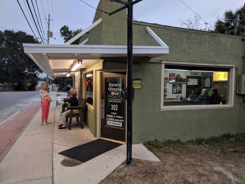 A small, green building with a sign for "Gene's Oyster Bar," featuring outdoor seating and patrons inside.
