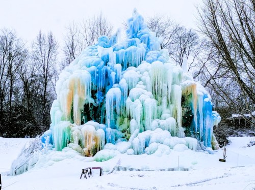A colorful ice formation resembling a frozen waterfall, surrounded by snow and bare trees in a winter landscape.