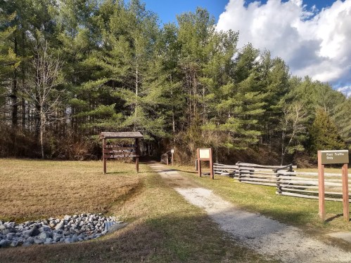 A gravel path leads into a wooded area, flanked by signs and a wooden fence under a blue sky with clouds.