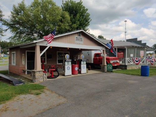 A vintage gas station with two pumps, flags, and a red truck parked outside, set in a small-town atmosphere.