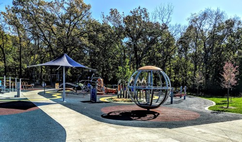A colorful playground with various equipment, including a large spinning structure, surrounded by trees and blue sky.