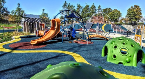 Colorful playground with slides, climbing structures, and soft rubber surfaces surrounded by trees.