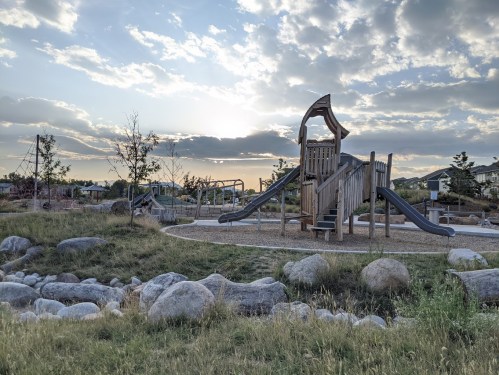 A playground with a wooden structure and slides, surrounded by grass and rocks, under a cloudy sky at sunset.