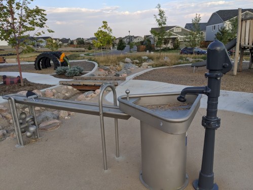 A playground water feature with a pump and a basin, surrounded by pathways and greenery.