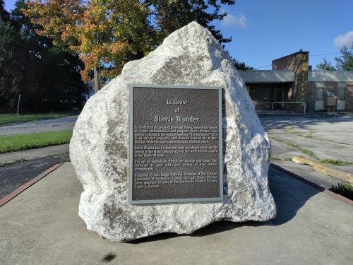 A large rock monument with a plaque honoring Stevie Wonder, set in an outdoor area with trees and a building in the background.
