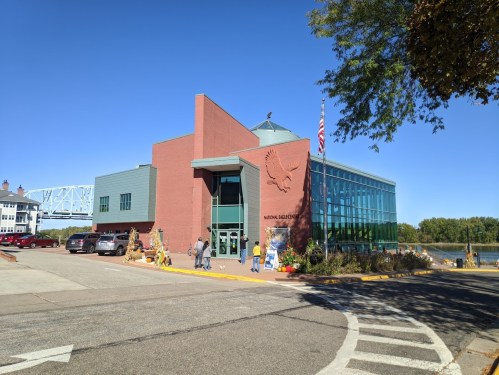 A modern building with large windows, featuring an eagle emblem, surrounded by trees and parked cars on a sunny day.