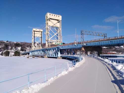 A blue bridge spans a frozen river, with snow-covered pathways and a clear blue sky in the background.