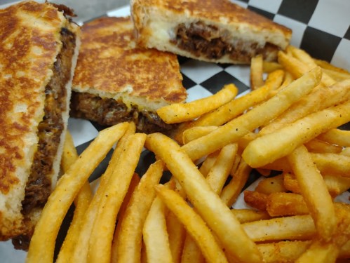 A close-up of a grilled sandwich filled with meat, served with a side of golden French fries on a checkered paper.