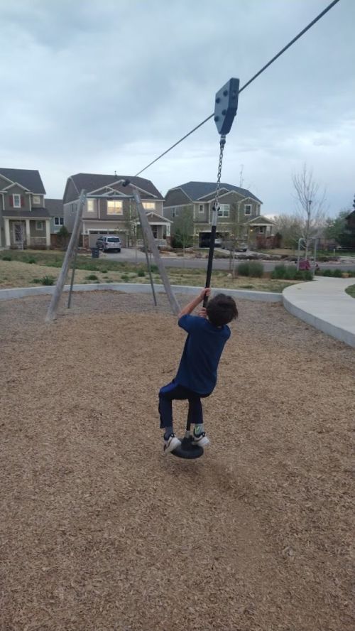 A child plays on a zip line at a park, holding onto a handle while suspended above a gravel surface.