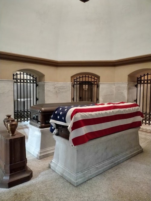 A casket draped with an American flag in a solemn, marble-floored mausoleum with decorative iron gates.