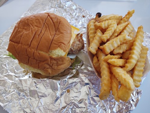 A crispy chicken sandwich with lettuce and cheese, served with a side of crinkle-cut fries on foil.