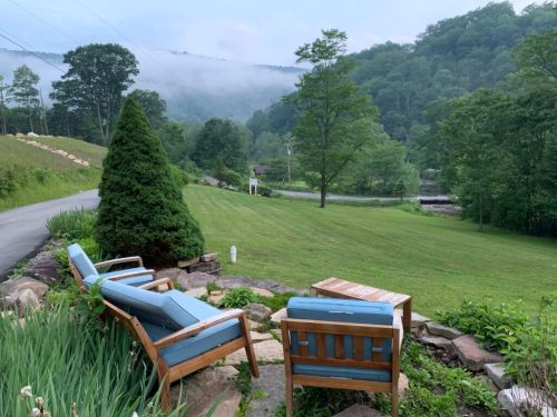 A serene outdoor scene with two blue chairs and a small table overlooking a green lawn and misty mountains.