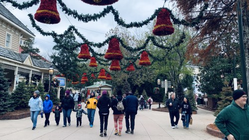 A festive street scene with people walking under decorated bell-shaped ornaments and holiday greenery.