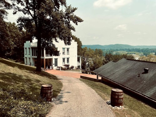 A scenic view of a white house on a hillside, with a gravel path and barrels, surrounded by trees and rolling hills.
