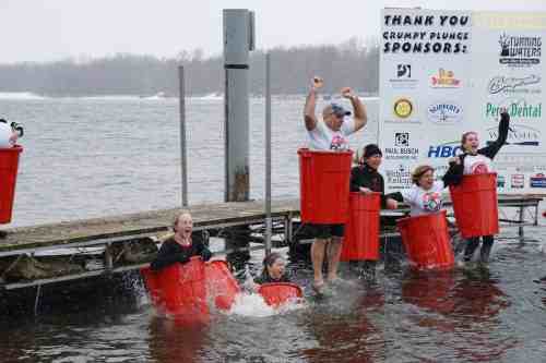 Participants joyfully plunge into icy water from red buckets, celebrating at a winter event by the dock.