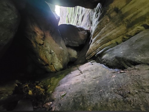 A narrow passage between large rocks, with sunlight illuminating the stone surfaces and scattered leaves on the ground.