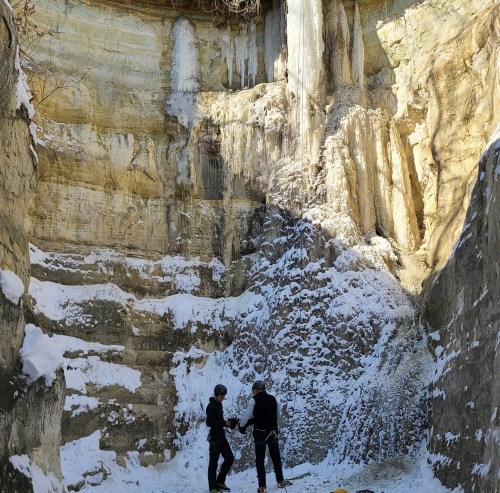 Two climbers prepare to ascend a snowy, icy rock face in a canyon surrounded by tall, frozen cliffs.