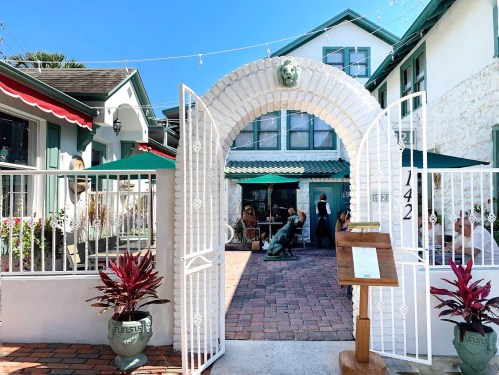 A white archway leads to a courtyard with potted plants, tables, and people enjoying the outdoor space.