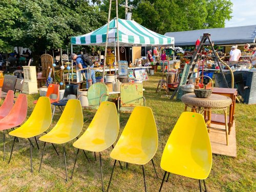A vibrant outdoor market scene featuring colorful chairs, various furniture, and a striped tent in the background.