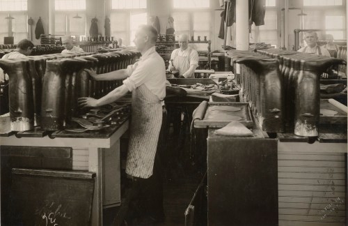 Workers in a shoe factory, focused on crafting boots, with rows of shoe molds in a well-lit workshop.