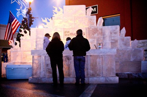 People gather around a large ice sculpture at dusk, with festive lights and an American flag in the background.