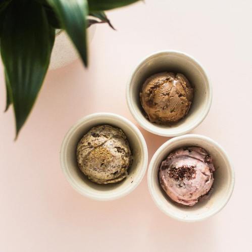 Three small bowls of ice cream in various flavors, placed on a light pink surface with a green plant in the background.