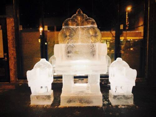 An ornate ice sculpture of a throne with two matching chairs, illuminated in a dark setting.