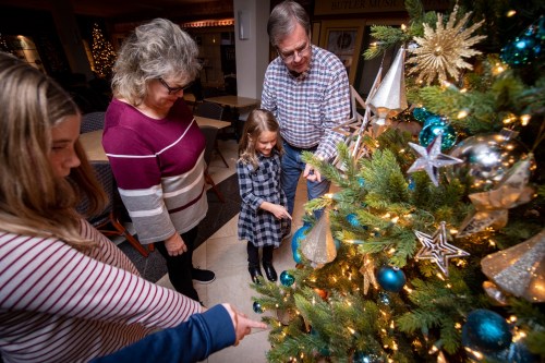 A family gathers around a decorated Christmas tree, pointing at ornaments and enjoying the festive atmosphere.