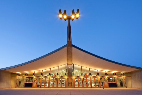 Modern building entrance with a distinctive roof design and tall light fixtures against a twilight sky.