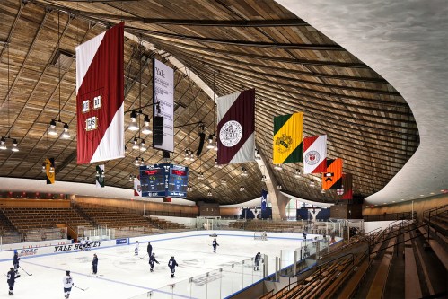 Interior of a hockey rink with wooden ceiling, flags hanging, and players on the ice. Spectator seating visible.