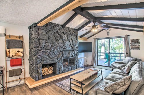 Cozy living room with a stone fireplace, wooden beams, and large windows overlooking a deck. Modern furnishings and decor.