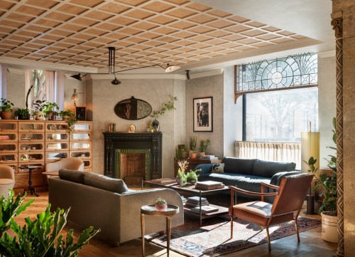 Cozy living room with plants, a fireplace, and large windows, featuring modern furniture and warm lighting.
