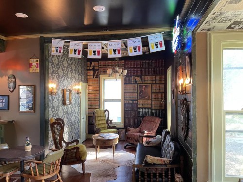 Cozy room with vintage decor, bookshelves, and a banner of drinks hanging from the ceiling. Soft lighting and seating areas.