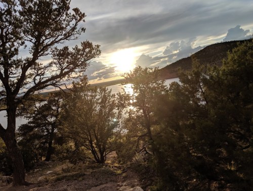 Sunset over a lake, framed by trees and mountains, with a cloudy sky reflecting warm light on the water.