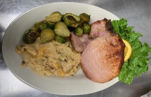 A plate with sliced ham, creamy mashed potatoes, Brussels sprouts, and a garnish of lettuce and lemon.