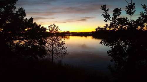 A serene sunset over a calm lake, framed by silhouettes of trees and a colorful sky.